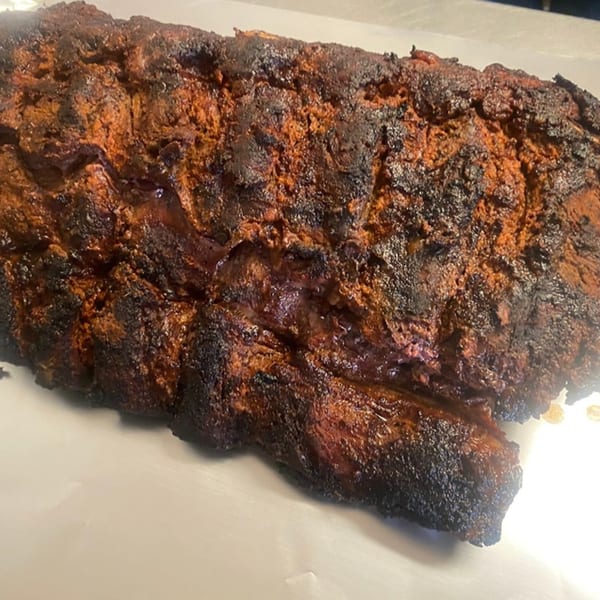 Close-up of a large, dark, smoky slab of BBQ meat with a rich, reddish-brown crust.