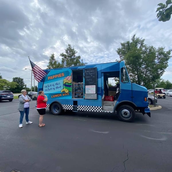 Two women stand near the blue Pappies Smokehouse & Lunch Box food truck with an American flag.